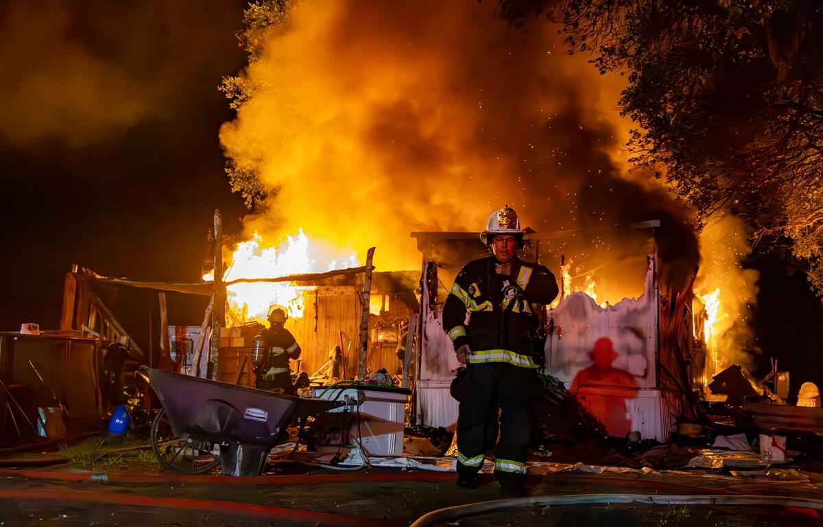 Firefighters work in front of a mobile home fully engulfed in flames at night, with thick smoke and burning debris surrounding the scene.