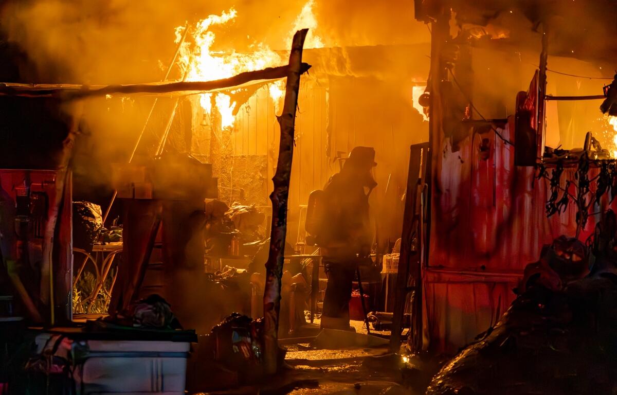 Silhouetted firefighter stands inside a smoke-filled, partially collapsed mobile home with flames pouring from the walls and roof.
