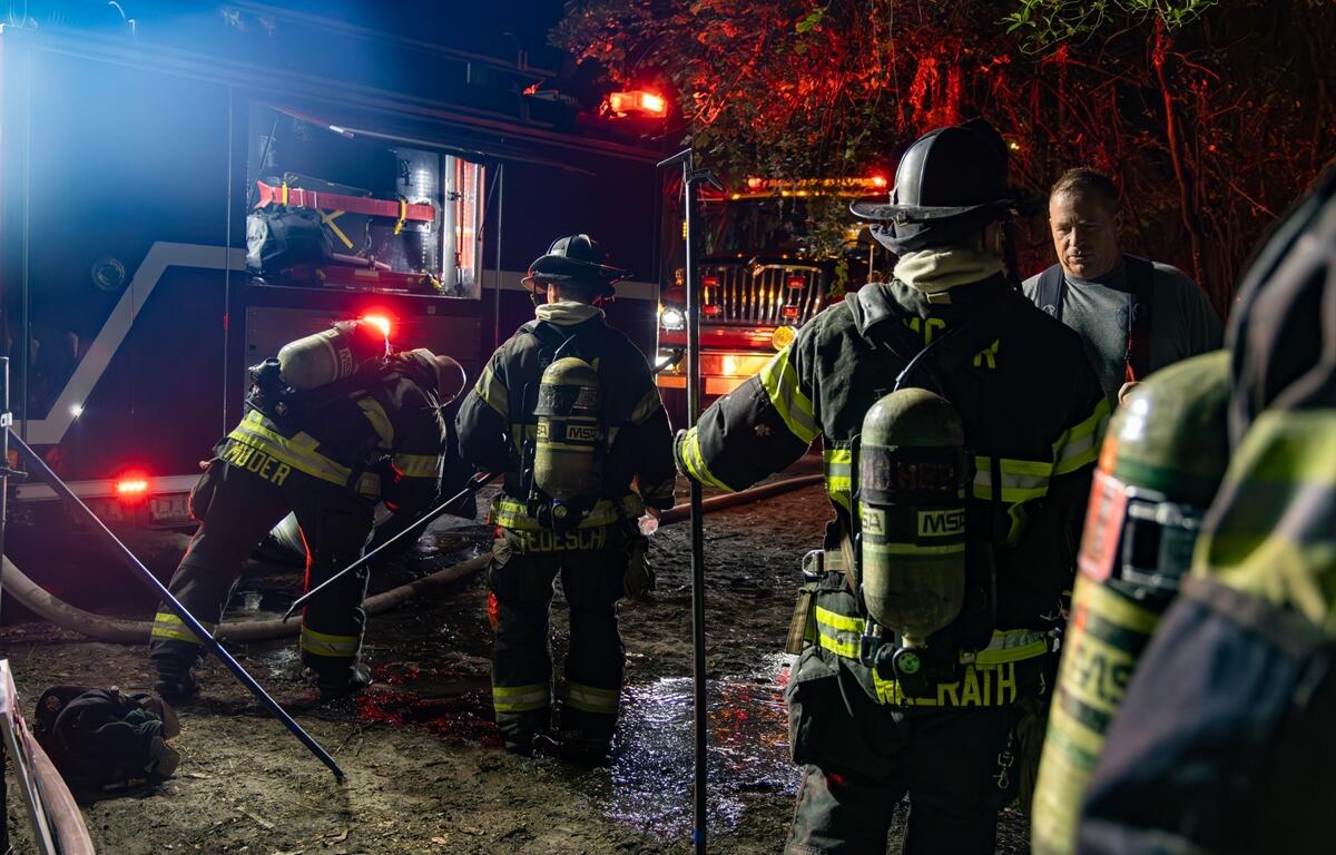Group of firefighters in full gear gather near a fire engine at night, with one firefighter crouched near open equipment and others preparing hoses.