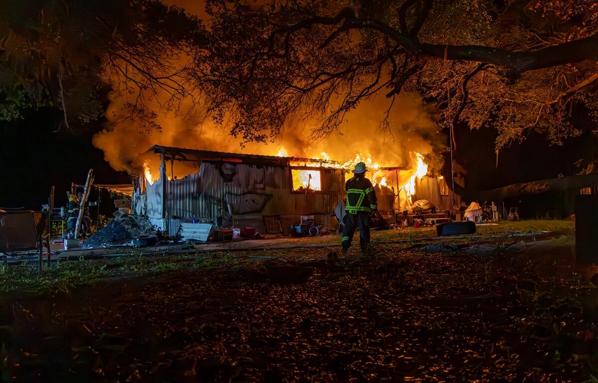 Firefighter walks toward a burning mobile home at night, with intense flames and smoke rising through the roof and trees silhouetted in the foreground.