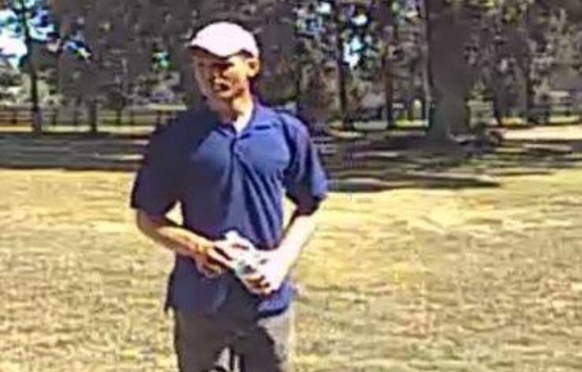 The photo shows a man standing on a grassy field with scattered trees in the background. He appears to be in his 20s or 30s, wearing a white baseball cap, a dark blue polo shirt, and dark pants. He is holding a white object—possibly a rolled-up shirt or towel—in both hands. The image is from surveillance footage and is slightly grainy, taken in daylight under clear skies.