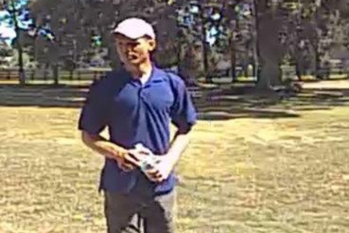 The photo shows a man standing on a grassy field with scattered trees in the background. He appears to be in his 20s or 30s, wearing a white baseball cap, a dark blue polo shirt, and dark pants. He is holding a white object—possibly a rolled-up shirt or towel—in both hands. The image is from surveillance footage and is slightly grainy, taken in daylight under clear skies.