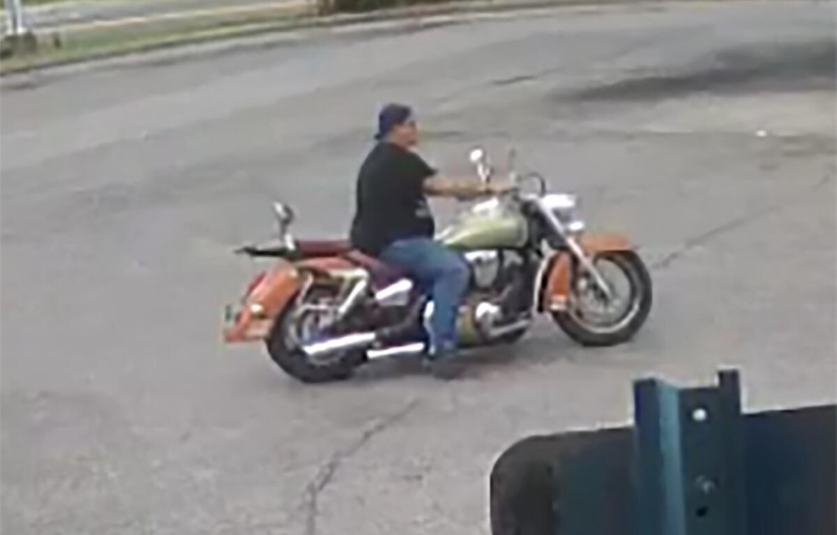 Accused Circle K thief wearing a black hat, black shirt, jeans, and boots rides an orange motorcycle.