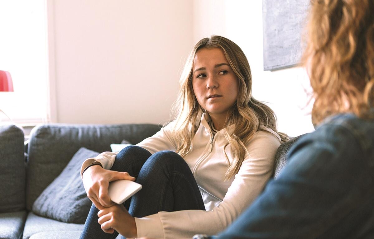 Young woman sitting on a couch, holding a phone and talking to another person during a serious conversation about mental health.