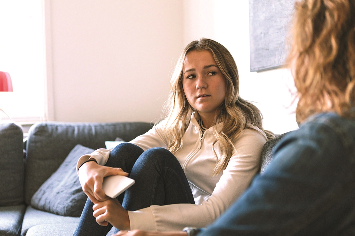 Young woman sitting on a couch, holding a phone and talking to another person during a serious conversation about mental health.