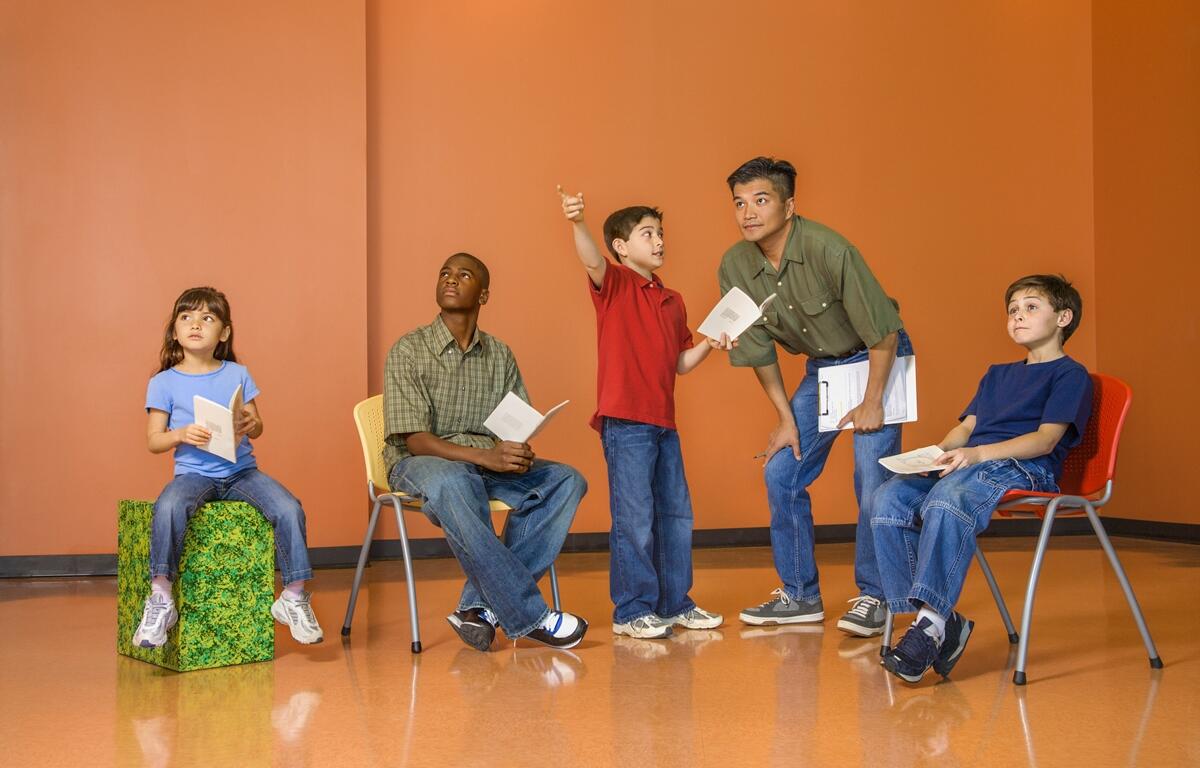Five children, including one adult or teen instructor, rehearse a dramatic scene in a classroom with orange walls. They hold scripts and appear engaged in acting or dialogue.
