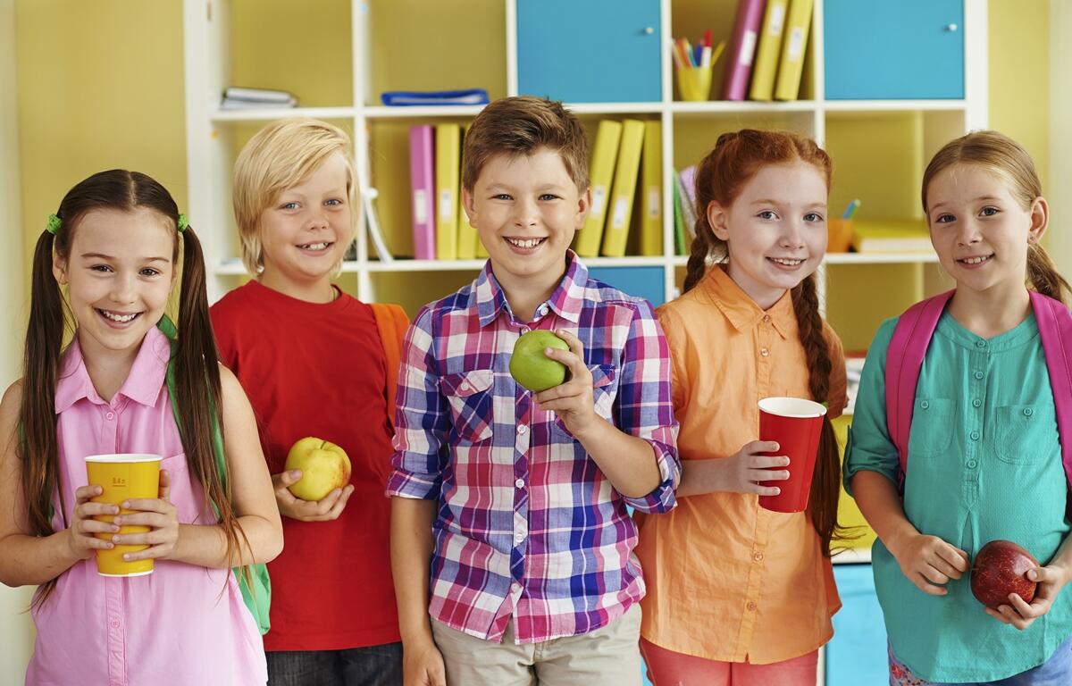 Five smiling children holding fruit and drinks during a summer library program, standing in front of colorful shelves.