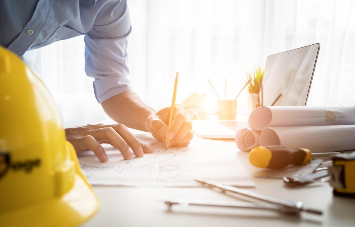 Person sketching building plans at a desk with blueprints, hard hat, laptop, and construction tools in daylight.