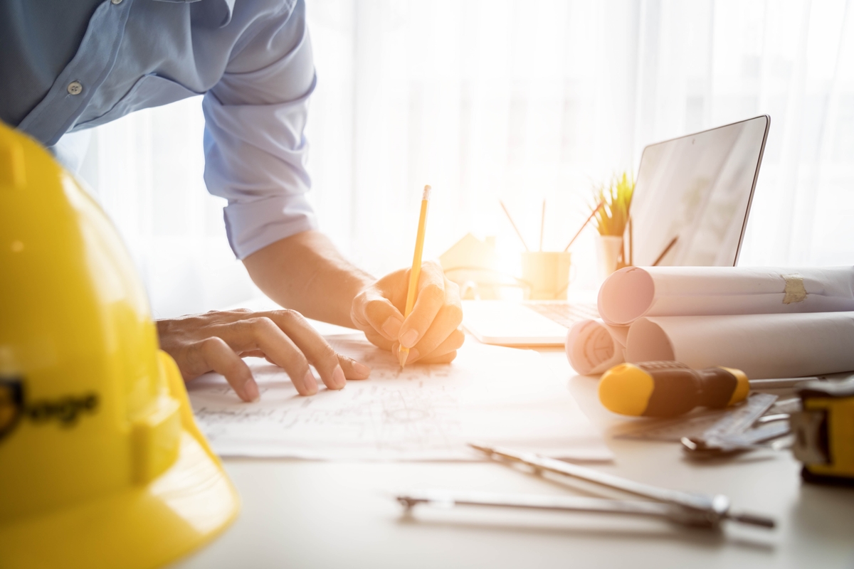 Person sketching building plans at a desk with blueprints, hard hat, laptop, and construction tools in daylight.