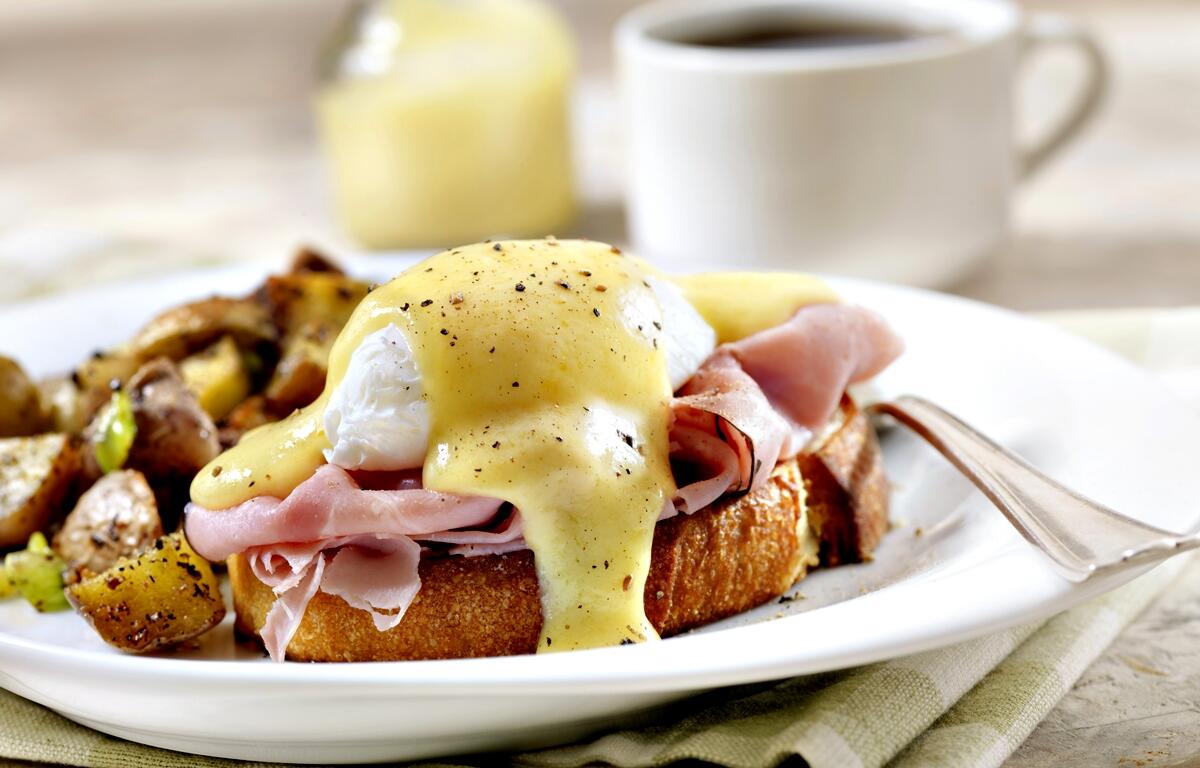 Plate of eggs Benedict with poached eggs, ham, and hollandaise sauce on toast, served with seasoned roasted potatoes and a cup of coffee in the background.