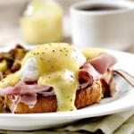 Plate of eggs Benedict with poached eggs, ham, and hollandaise sauce on toast, served with seasoned roasted potatoes and a cup of coffee in the background.