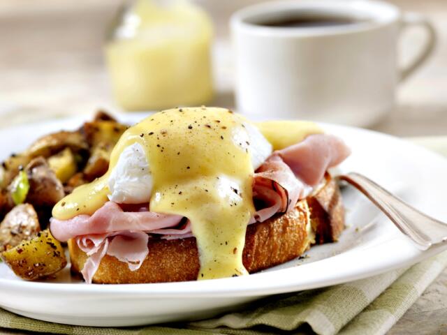 Plate of eggs Benedict with poached eggs, ham, and hollandaise sauce on toast, served with seasoned roasted potatoes and a cup of coffee in the background.