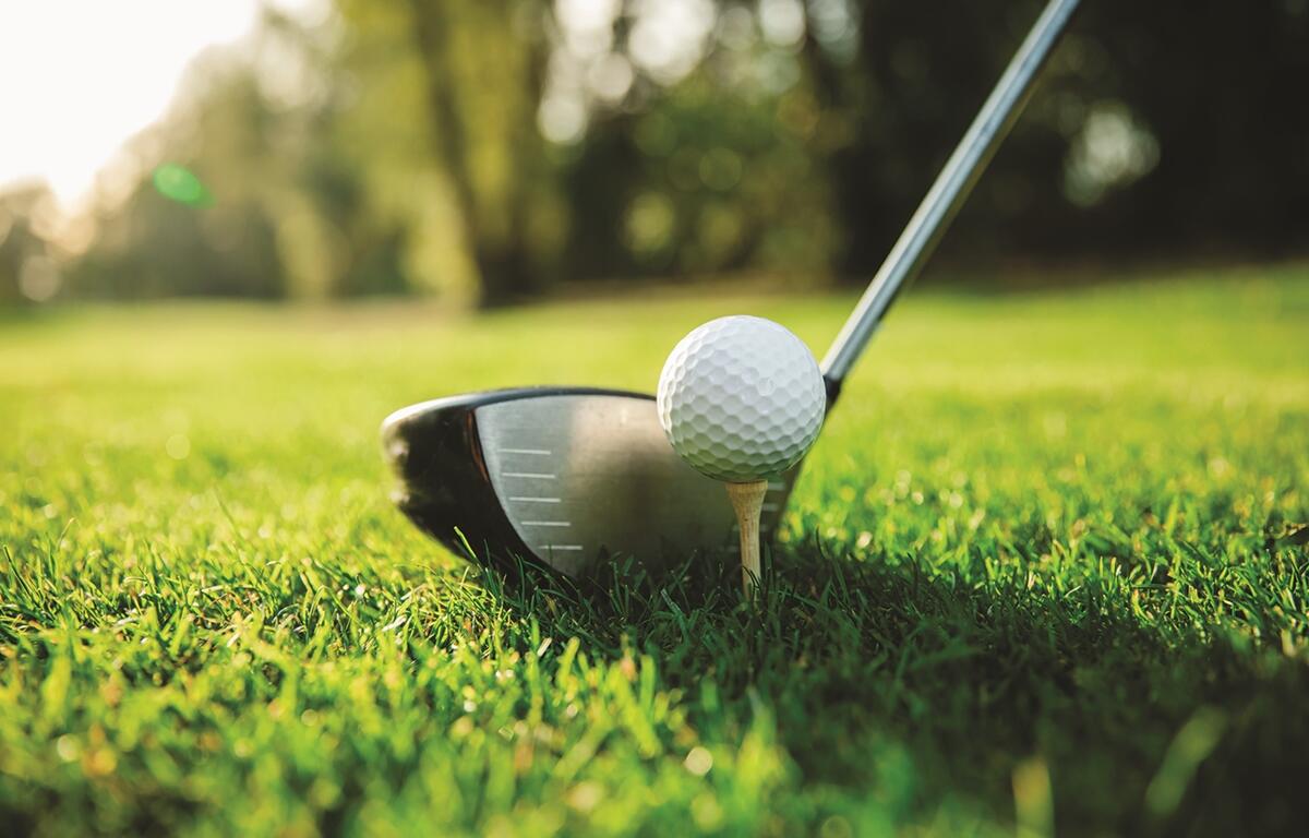 Close-up of a golf club about to strike a golf ball on a tee with green grass in the background.