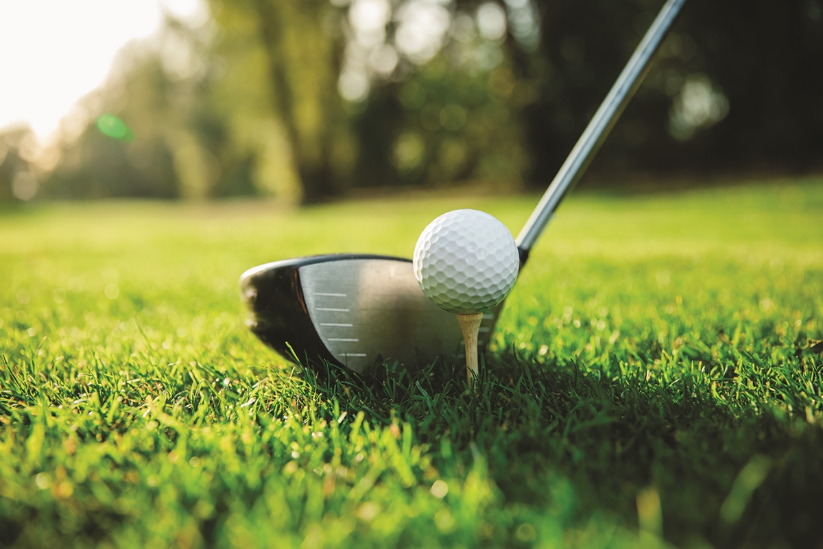 Close-up of a golf club about to strike a golf ball on a tee with green grass in the background.