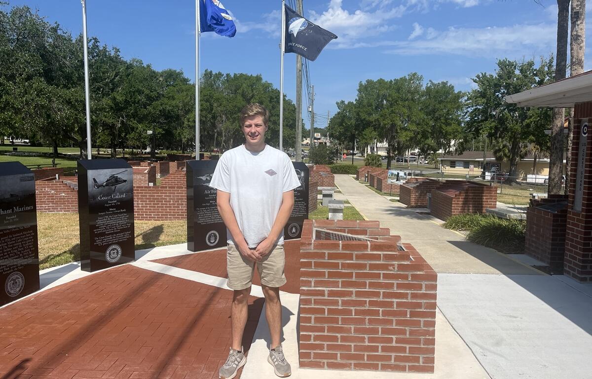 A young man stands in front of a red brick and marble military flag display at the Ocala/Marion County Veterans Memorial Park, with flagpoles and engraved memorial slabs visible behind him under a sunny sky.