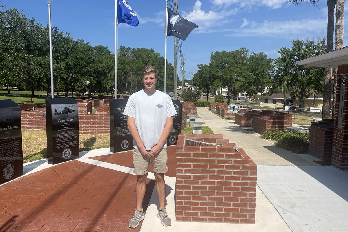 A young man stands in front of a red brick and marble military flag display at the Ocala/Marion County Veterans Memorial Park, with flagpoles and engraved memorial slabs visible behind him under a sunny sky.