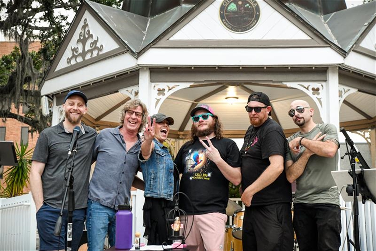 Heather Lynne stands with her band in front of a white gazebo, smiling and making hand gestures after a live performance in downtown Ocala.