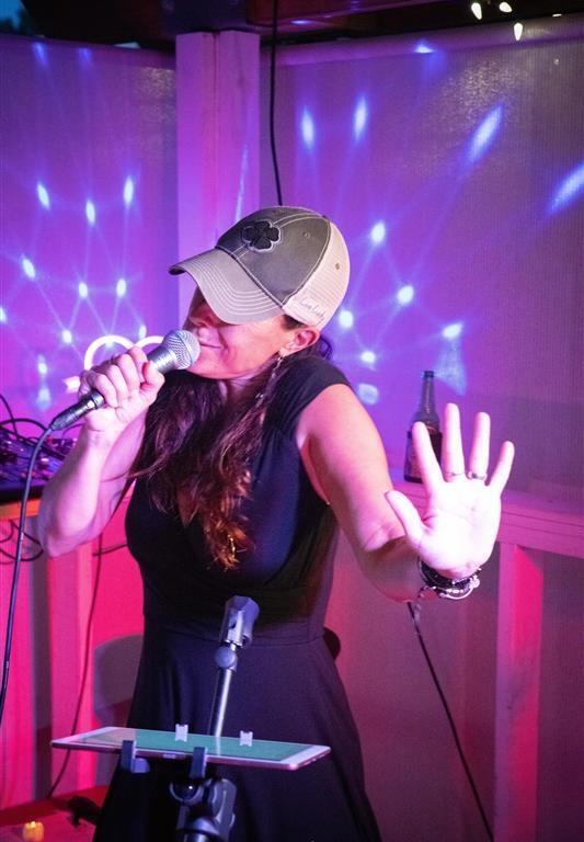 Heather Lynne sings into a microphone with her hand raised, wearing a casual ball cap and black dress under vibrant purple and blue stage lighting.