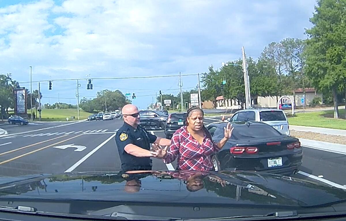 Ocala Police officers arrest a woman next to a silver vehicle on SW College Road following a reported bank robbery; the scene includes police vehicles and flashing lights.