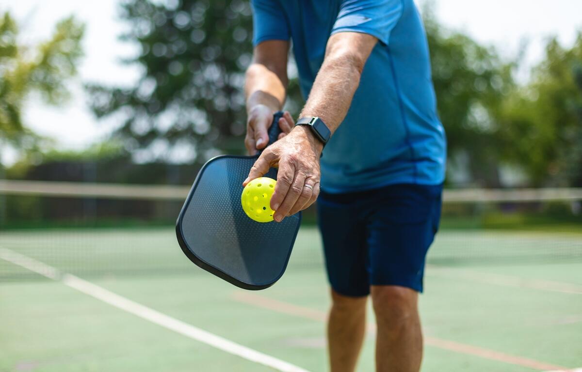 Close-up of a man in a blue shirt and shorts getting ready to serve a yellow pickleball with a black paddle on an outdoor court.