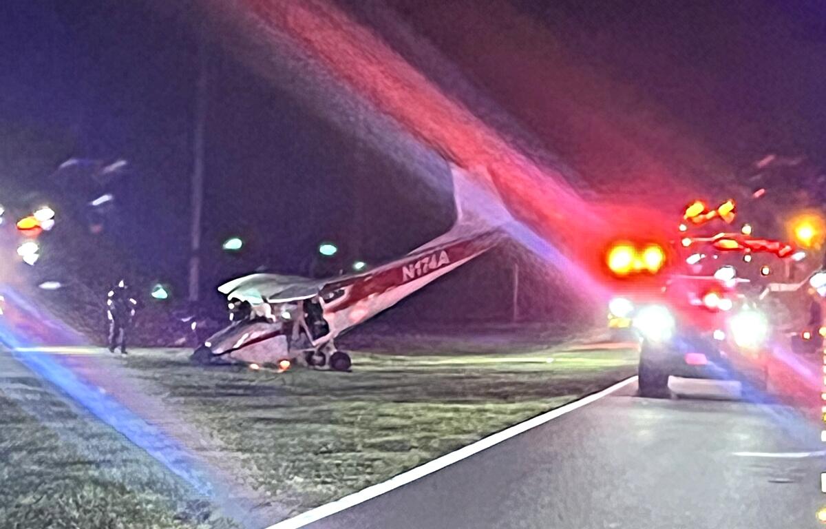 A small Cessna aircraft sits damaged in the grass near a roadway at night, surrounded by emergency vehicles with flashing lights, as first responders assess the crash scene in Ocala, Florida.