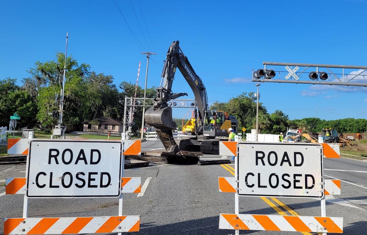 Two "Road Closed" barricades block the entrance to a railroad crossing on U.S. 301 in Wildwood, Florida, where workers and an excavator are actively removing railroad tracks under clear blue skies.