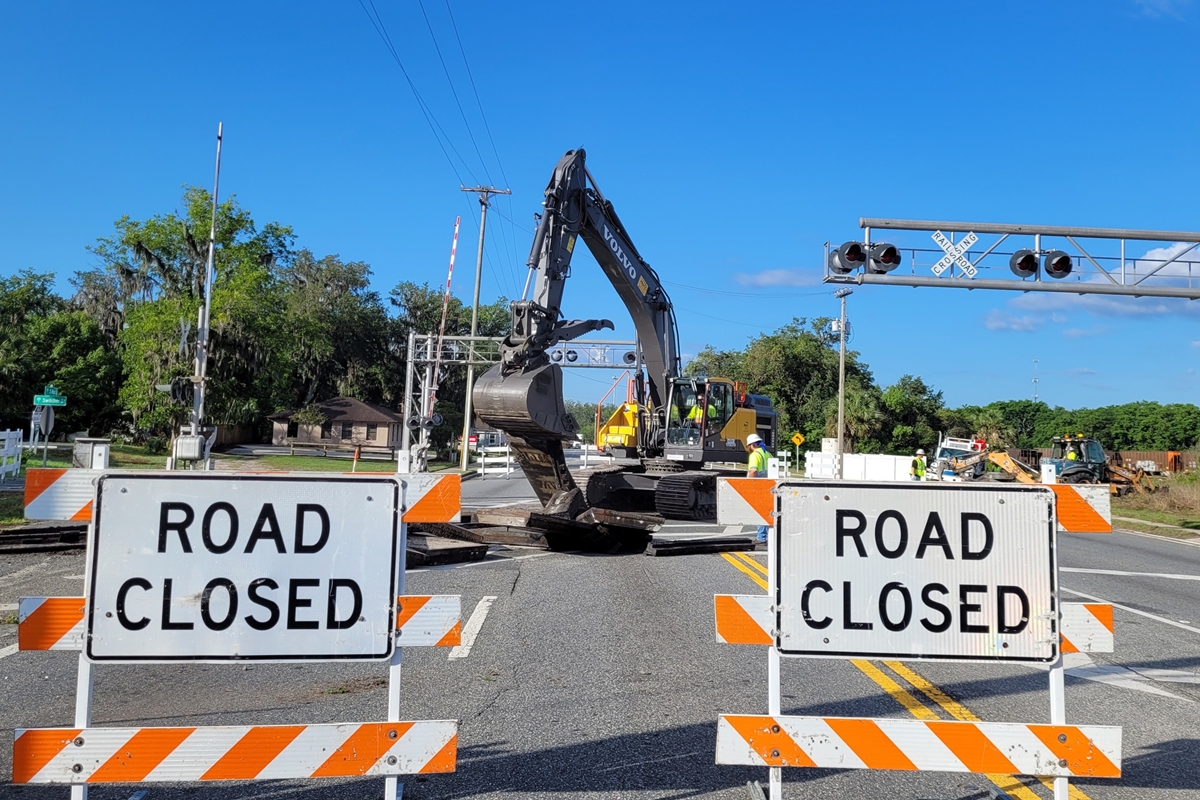Two "Road Closed" barricades block the entrance to a railroad crossing on U.S. 301 in Wildwood, Florida, where workers and an excavator are actively removing railroad tracks under clear blue skies.