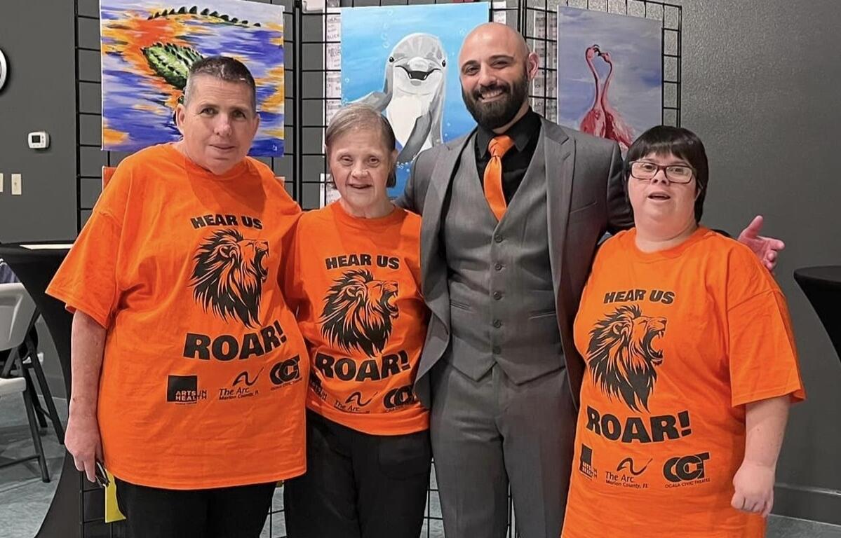 Three Arc of Marion clients wearing matching orange "Hear Us Roar" shirts stand with AIHOM artist Christopher Hershberger in front of colorful animal paintings.