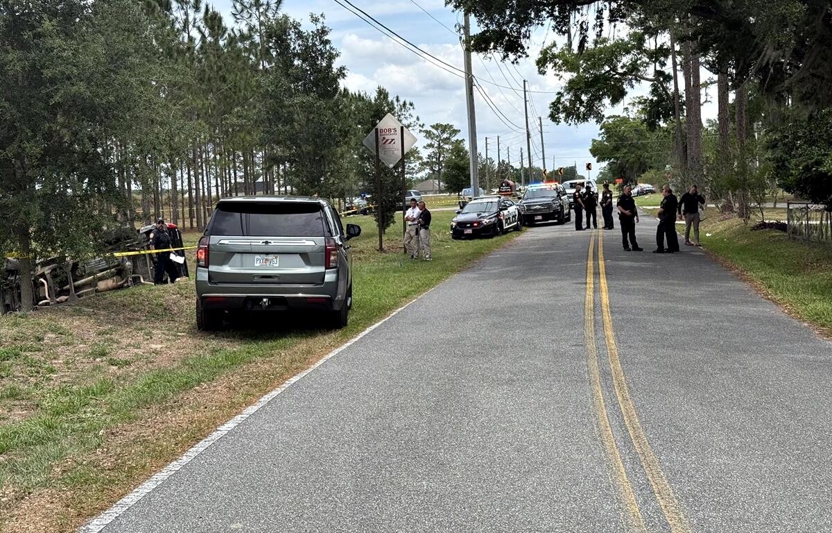 Law enforcement officers and vehicles line a two-lane road near a wooded area where an overturned vehicle is visible behind yellow crime scene tape.
