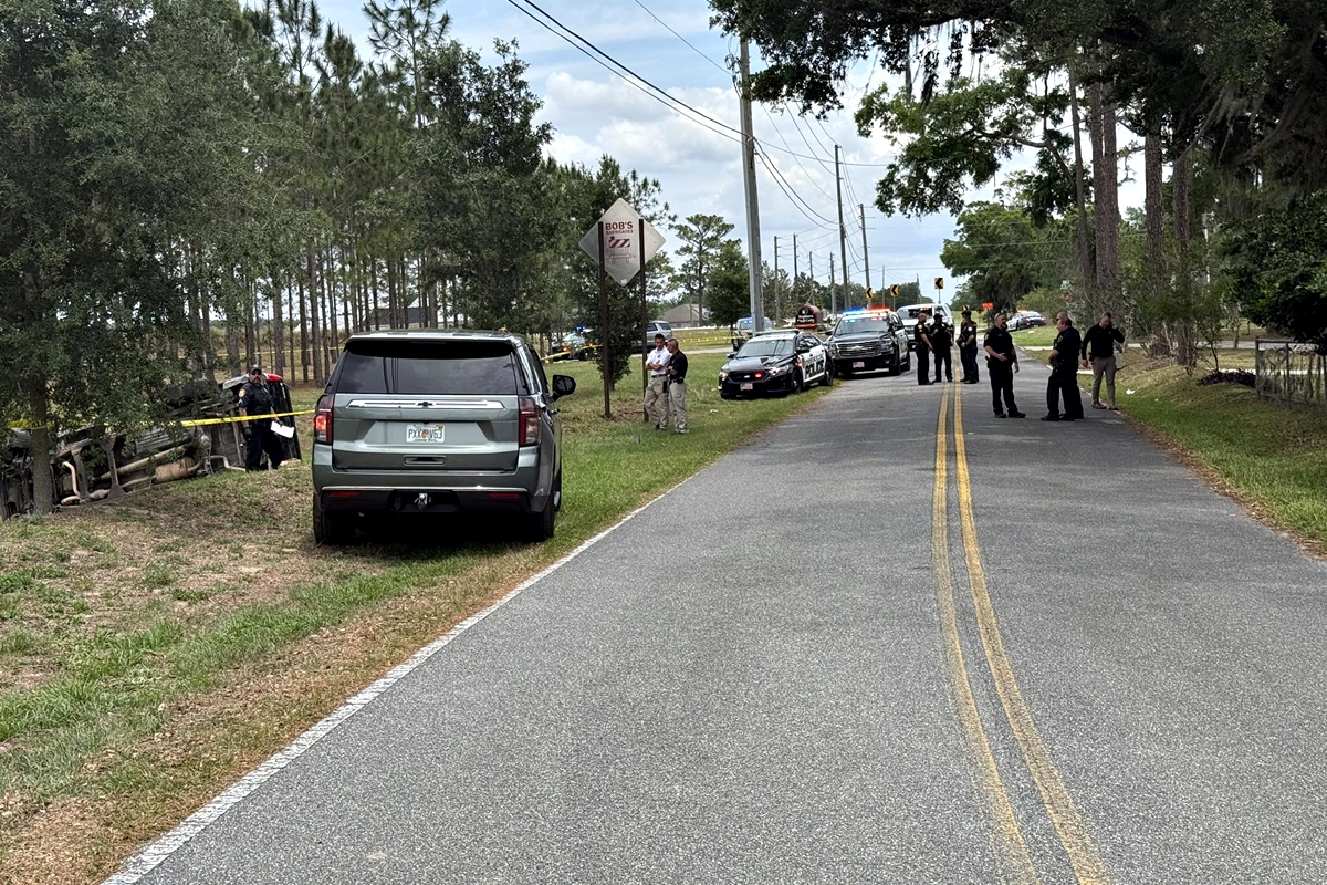 Law enforcement officers and vehicles line a two-lane road near a wooded area where an overturned vehicle is visible behind yellow crime scene tape.