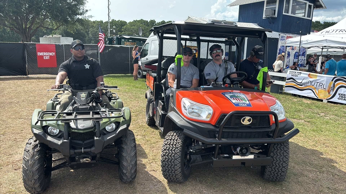 A sheriff's deputy on an ATV and a group of emergency responders in an orange utility vehicle are stationed near the emergency exit at the Rock the Country 2025 festival, with event tents, American flags, and sponsor signage visible in the background.