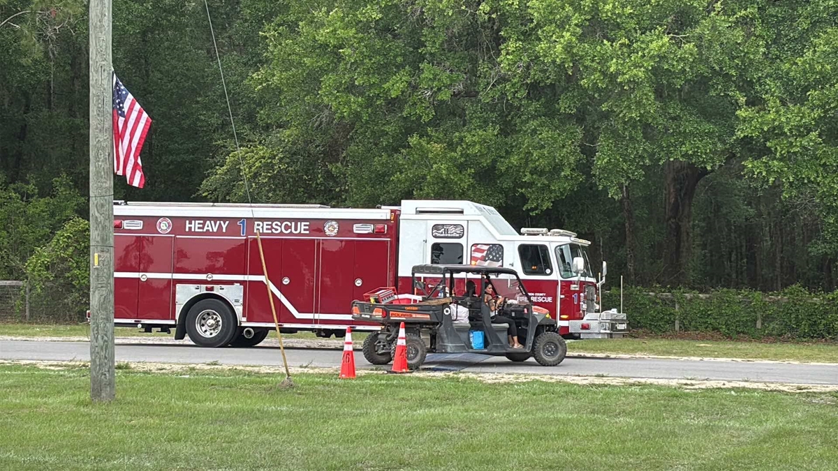 A Marion County Fire Rescue heavy rescue truck is parked along a tree-lined road beside a utility vehicle marked for fire rescue use. Two people are seated in the utility vehicle near orange traffic cones. In the foreground, an American flag hangs at half-staff from a wooden pole, and grassy open space surrounds the scene.