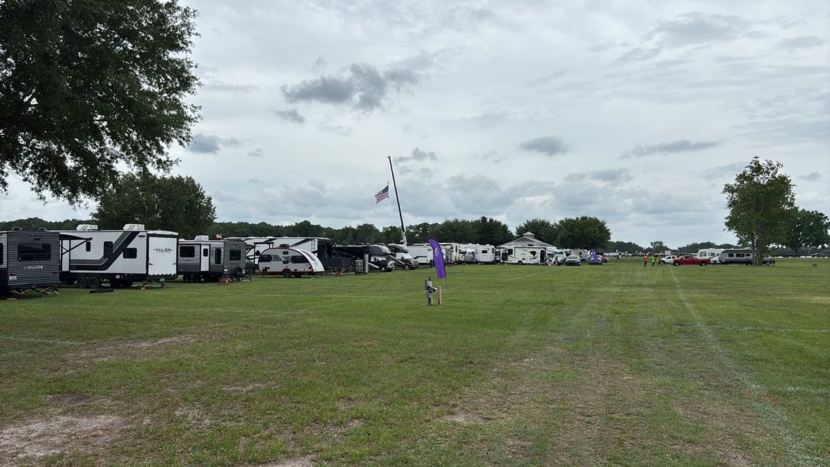 A row of RVs lines the edge of a wide grassy field under a cloudy sky at a campground area for the "Rock the Country" festival. An American flag hangs at half-staff from a tall crane in the distance. A few people and vehicles are scattered along the horizon, and a purple event marker stands in the foreground near electrical hookups.