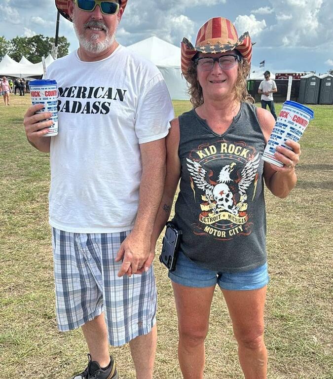 A man and woman smile while holding Rock the Country souvenir cups, wearing matching American flag cowboy hats and patriotic shirts—his reading “American Badass” and hers featuring Kid Rock. They stand on grass with event tents, portable toilets, and a crane-hoisted U.S. flag in the background under partly cloudy skies.