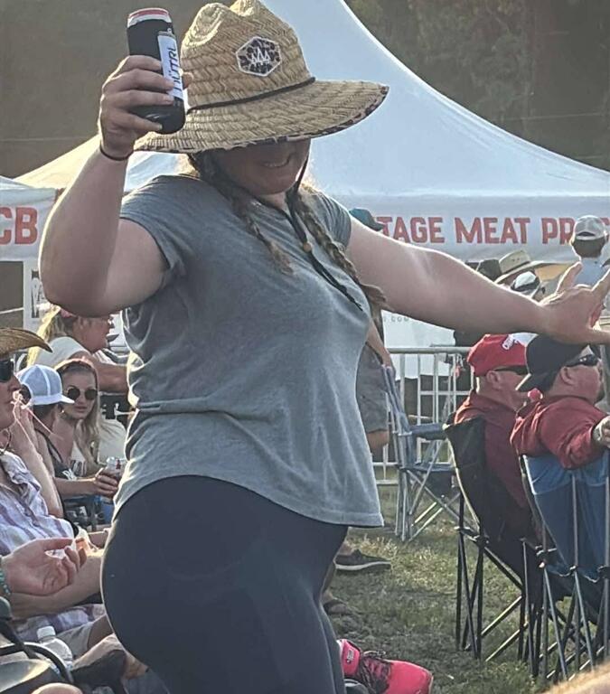 A woman in a wide-brimmed straw hat with braids smiles and raises a can of Busch Light while dancing at an outdoor festival. She wears a gray T-shirt and black leggings, surrounded by people seated in camping chairs near white vendor tents under the afternoon sun. The scene captures a lighthearted, celebratory moment.