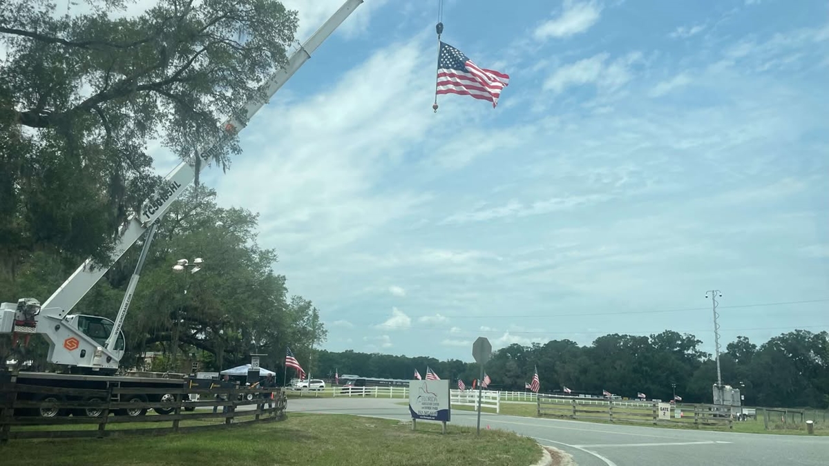 A large American flag hangs from a crane at the entrance to the Rock the Country festival at the Florida Horse Park, with smaller flags lining the fence and a directional sign pointing toward Gate 2 under a partly cloudy sky.