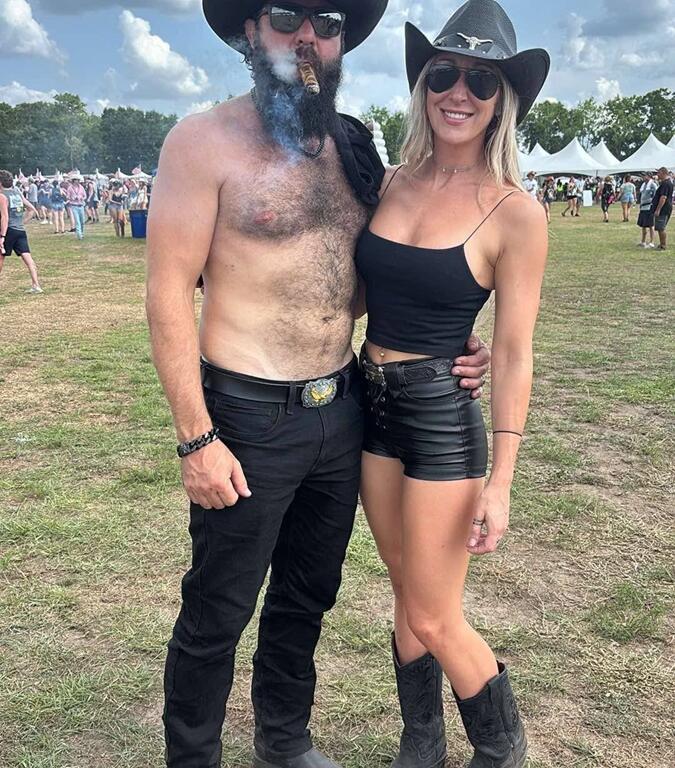 A man and woman pose together at an outdoor event, both wearing black cowboy hats and sunglasses. The man is shirtless, wearing black jeans and boots, with a cigar in his mouth and smoke rising. The woman wears a black crop top, high-waisted black shorts, and cowboy boots. Tents and festivalgoers can be seen in the background under a partly cloudy sky.