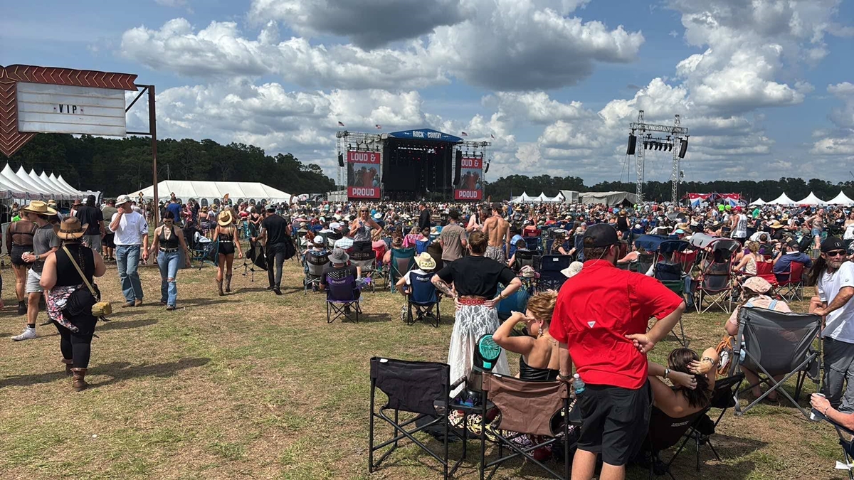 A large crowd gathers at the Rock the Country music festival under a partly cloudy sky, with attendees seated in camp chairs and walking toward the main stage, which is framed by banners reading “LOUD & PROUD.” White tents and a VIP sign are visible on the festival grounds.