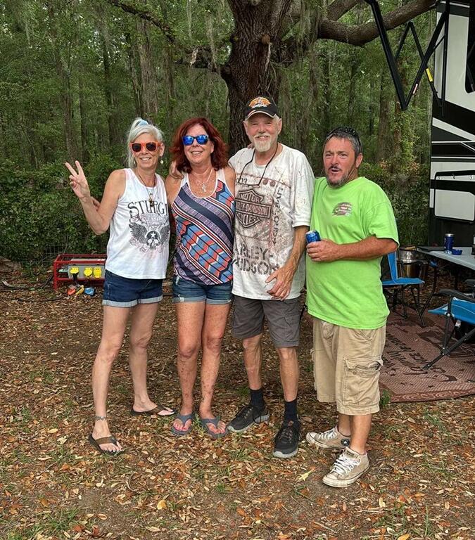 Four adults stand smiling under a large oak tree with Spanish moss, next to a camper at a wooded campsite. Two women on the left wear shorts, sunglasses, and tank tops—one flashing a peace sign—while the two men on the right, one in a Harley-Davidson shirt, have arms around each other and hold drinks. A small table, chairs, and generator are visible in the background.