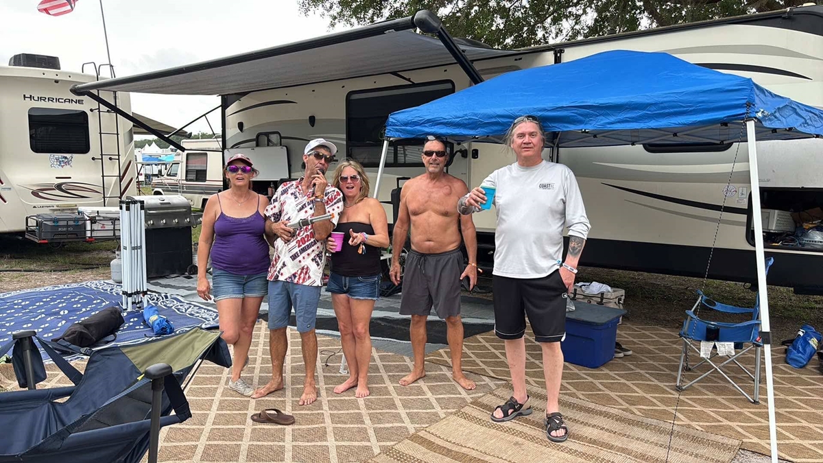 A group of five festivalgoers pose with drinks in hand at their RV campsite at Rock the Country, standing under an awning and pop-up tent with grills, chairs, and coolers set up on rugs outside their campers.