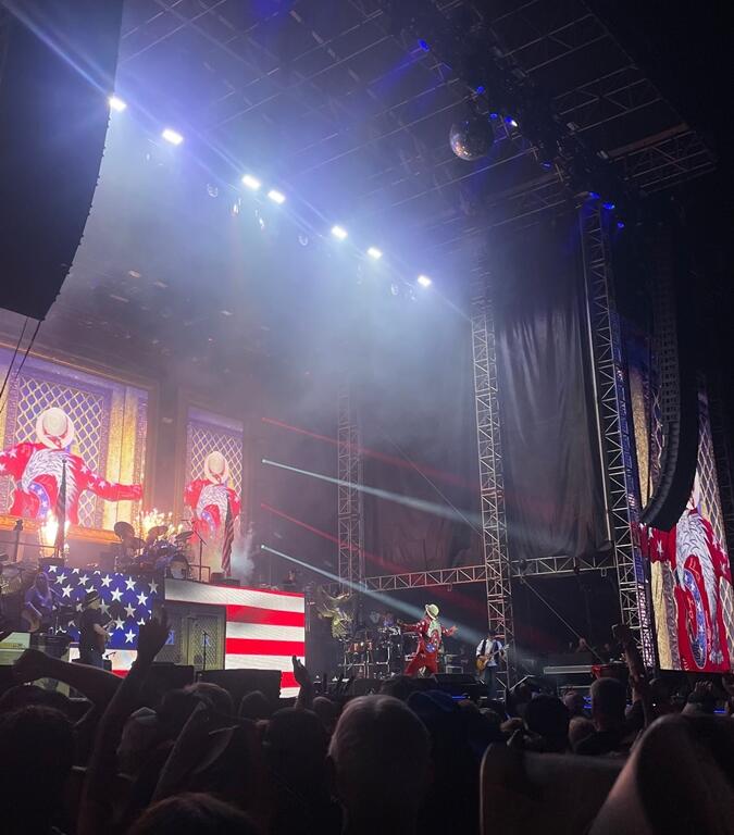 A vibrant concert scene at "Rock the Country" features a performer in a red, white, and blue outfit with a cowboy hat standing center stage, with American flag visuals and flames decorating the backdrop. Large screens show close-ups of the artist striking a dynamic pose. Bright lights beam across the stage as the crowd cheers and takes photos, many wearing cowboy hats in a patriotic atmosphere.