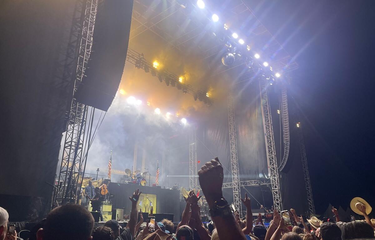 A lively crowd cheers with raised hands and cowboy hats as bright white and gold lights beam down from the "Rock the Country" concert stage. The stage is filled with smoke and lit dramatically, with American flags flanking the drum kit and instruments on stage. Fans near the front capture the moment on their phones, enjoying the high-energy nighttime performance.