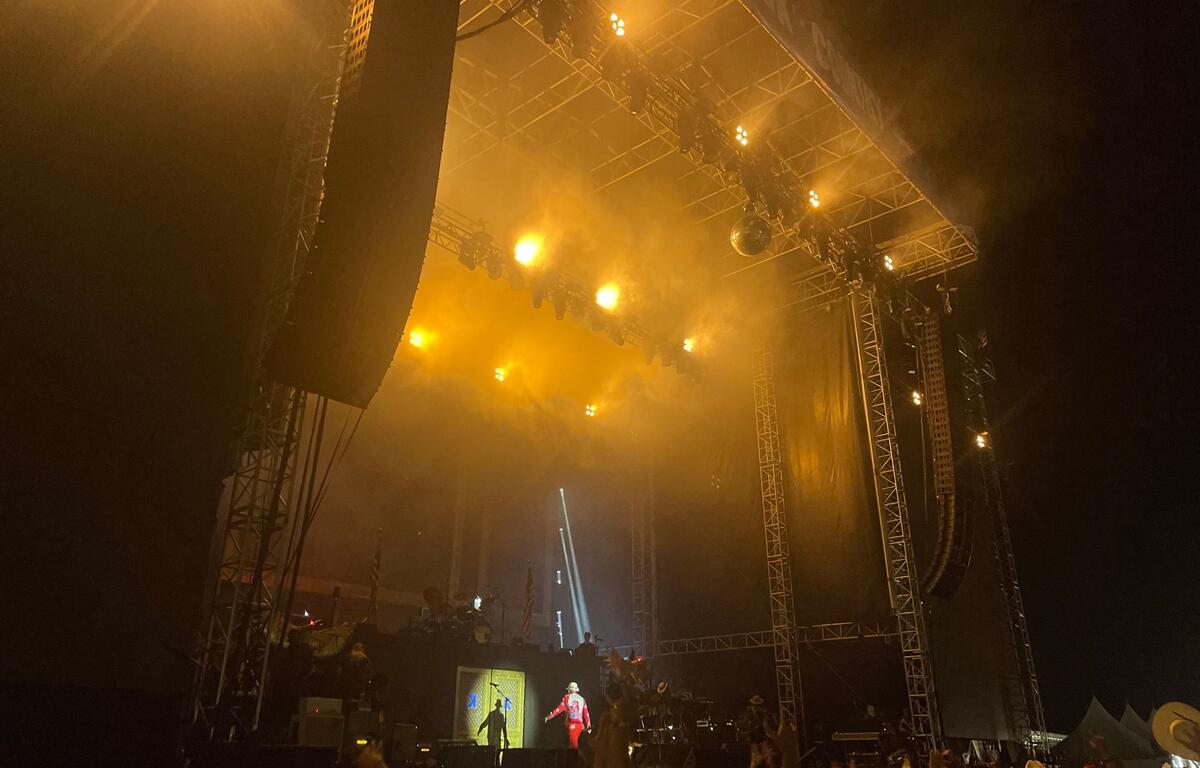 A performer dressed in red takes the stage under bright golden-yellow lights at the "Rock the Country" concert, with beams of light cutting through stage smoke. The crowd below cheers and raises their hands, many wearing cowboy hats. The large stage structure is lit dramatically against the dark night sky, creating an energetic and immersive concert atmosphere.