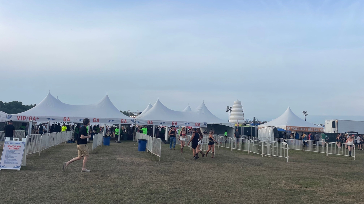 Attendees enter the "Rock the Country" music festival through a series of white canopy tents labeled GA (General Admission), GA+, and VIP. Metal barricades guide foot traffic, and event staff in neon green shirts manage the entry points. A sign lists items not allowed inside, while a nearby tent marked "Ticket Help / Will Call" serves waiting guests. In the background, a large inflatable ice cream cone rises near food trucks under a clear evening sky.
