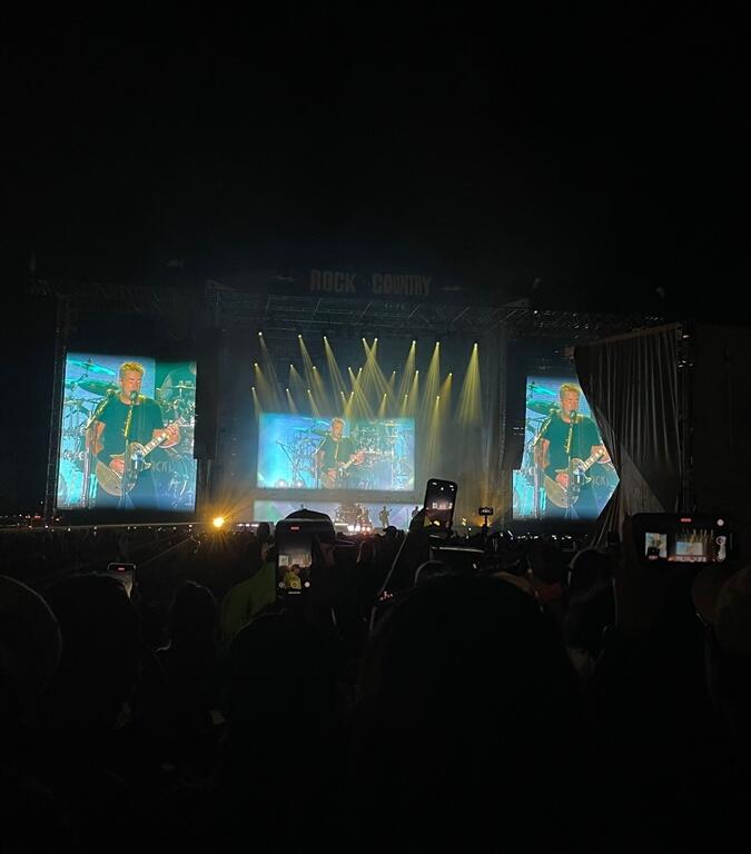 A nighttime crowd watches a live performance at the "Rock the Country" music festival. The stage is brightly lit with yellow beams and large video screens showing a musician singing and playing guitar. Audience members hold up phones to record the moment, silhouetted against the glow of the stage lighting and visuals.