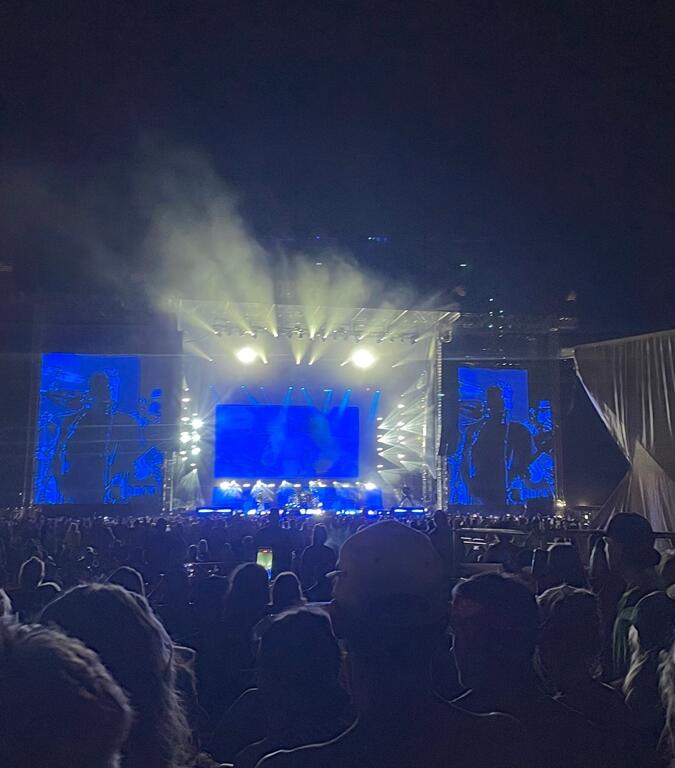 A large nighttime crowd watches a live performance at the "Rock the Country" festival. The stage is brightly lit with blue, yellow, and orange lights, and two giant video screens display a guitarist and singer in action. Audience members are seated and standing in the viewing area, silhouetted against the illuminated stage as music fills the outdoor venue.