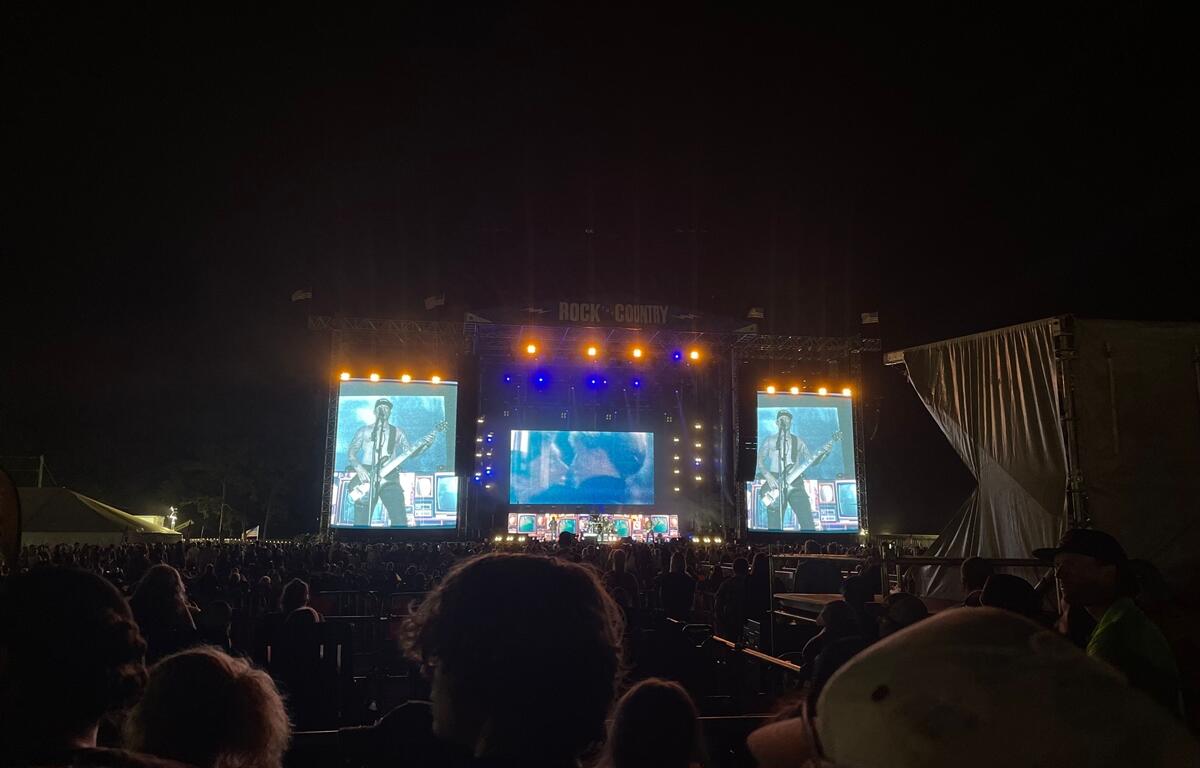 A large nighttime crowd watches a live performance at the "Rock the Country" festival. The stage is brightly lit with blue, yellow, and orange lights, and two giant video screens display a guitarist and singer in action. Audience members are seated and standing in the viewing area, silhouetted against the illuminated stage as music fills the outdoor venue.