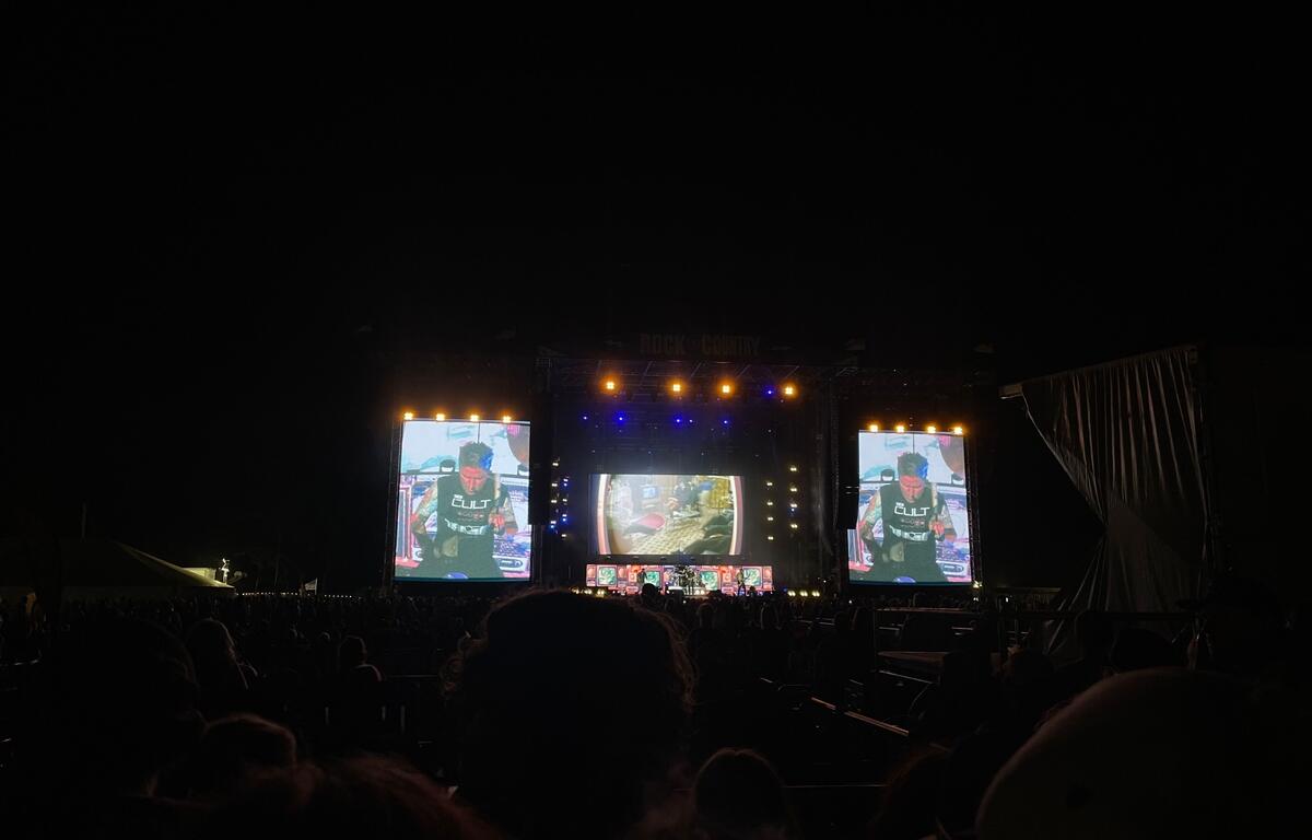 A nighttime crowd watches a live performance at the "Rock the Country" music festival. The stage is lit with orange and blue lights, and two large video screens display a close-up of the performer singing. Concertgoers fill the viewing area, standing or sitting in seats, as the energetic atmosphere builds under the dark sky.
