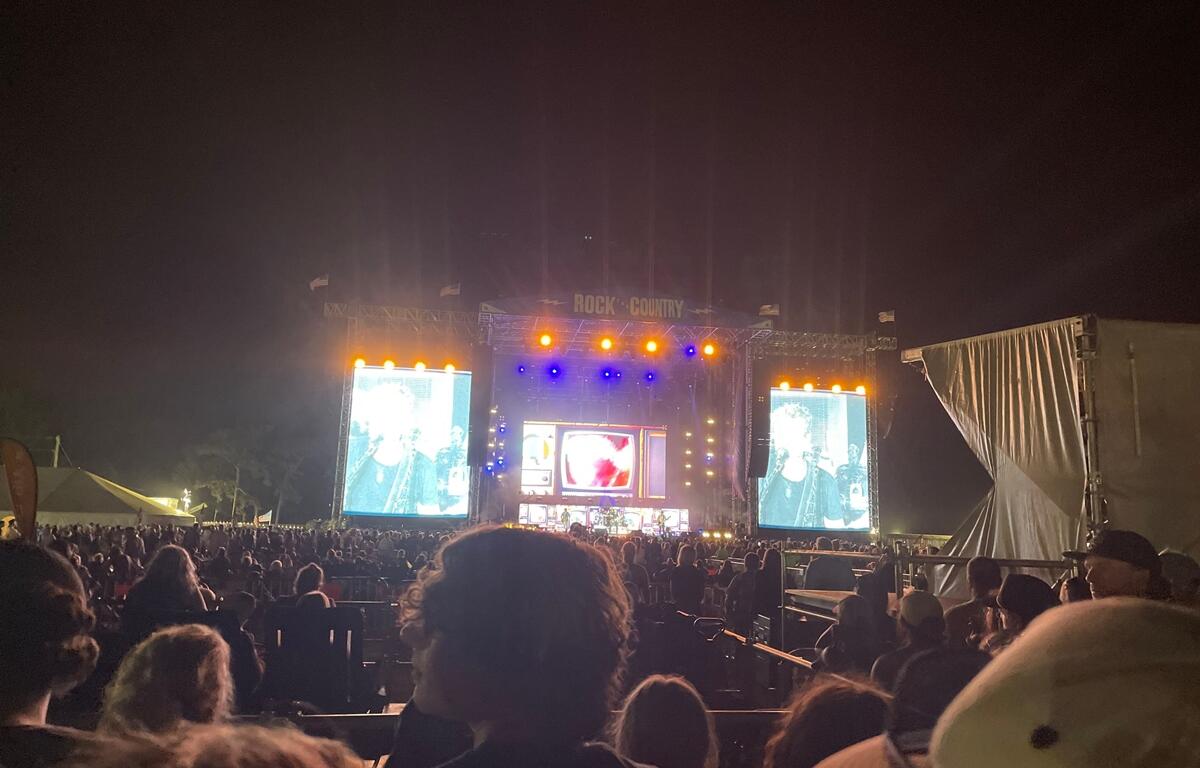 A nighttime crowd watches a live performance at the "Rock the Country" music festival. The stage is lit with orange and blue lights, and two large video screens display a close-up of the performer singing. Concertgoers fill the viewing area, standing or sitting in seats, as the energetic atmosphere builds under the dark sky.
