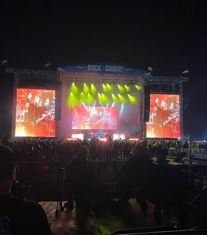 A large crowd enjoys a nighttime concert at the "Rock the Country" festival. Bright yellow stage lights beam outward as a performer sings on stage, shown on two massive video screens flanking the stage. The audience, some seated and some standing, is silhouetted in front of the brightly lit performance area under a clear, dark sky.
